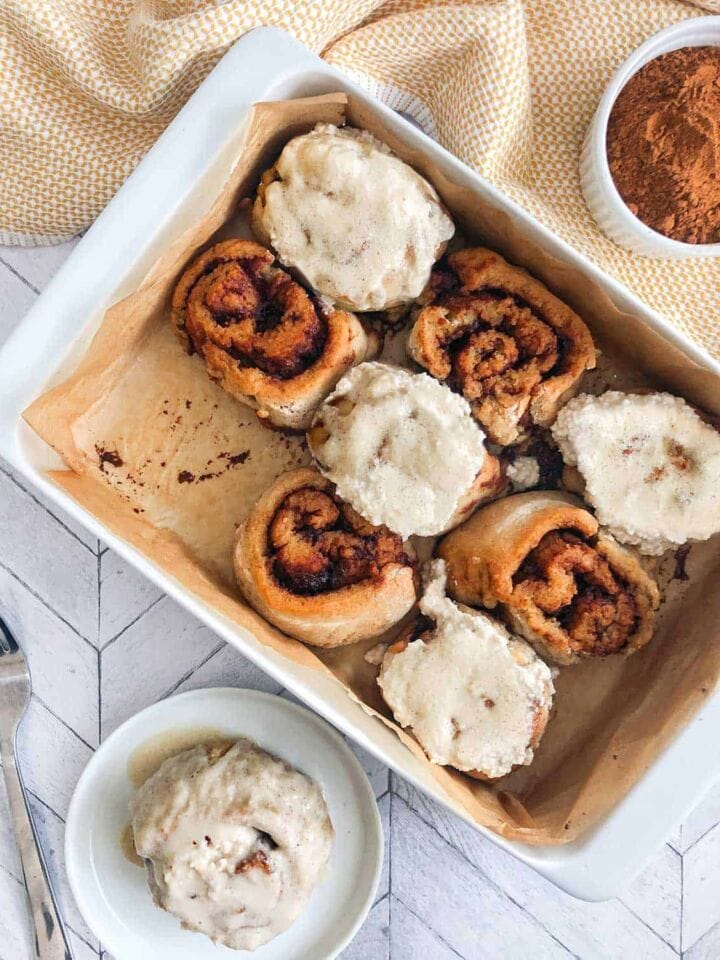 Vegan and Gluten Free Cinnamon Rolls with Maple Cashew Cream Cheese Icing in white baking dish with one roll in small plate with icing