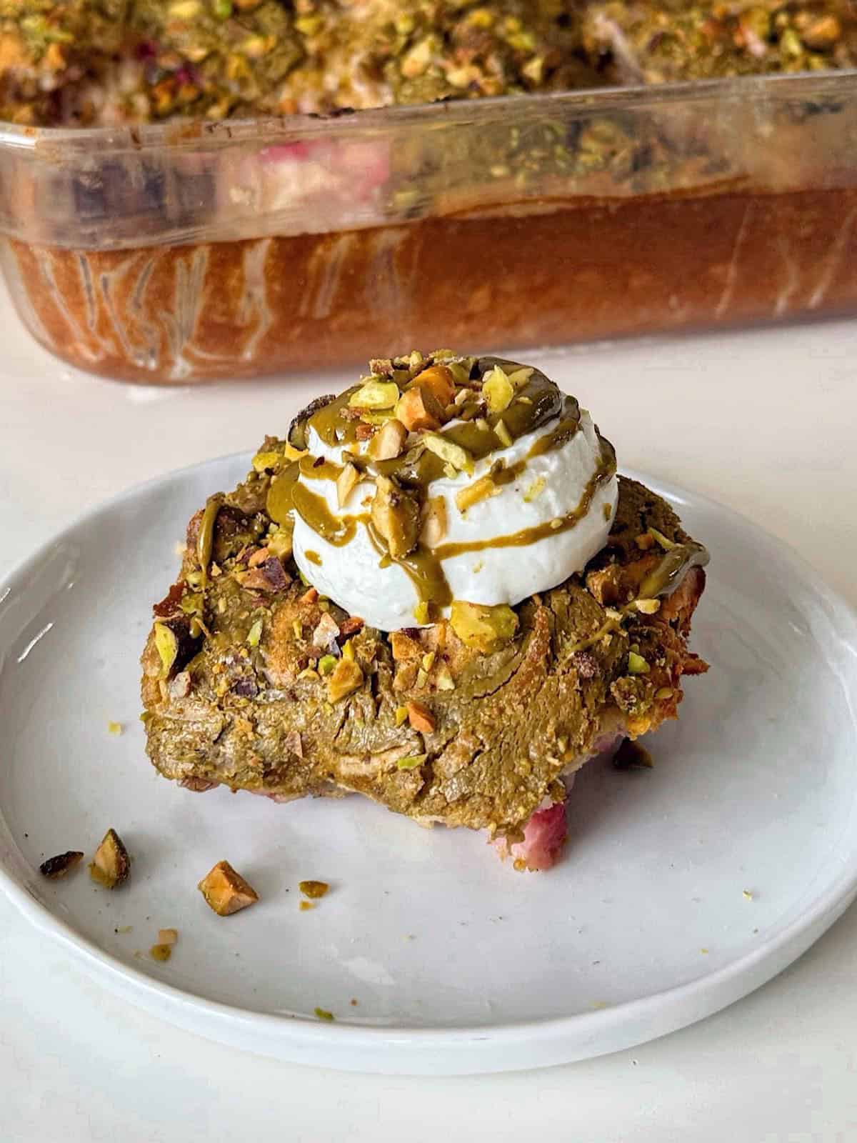 Strawberry Pistachio Baked Oats 1 Slice of strawberry pistachio baked oats topped with a dollop of yogurt, pistachio butter drizzle, and chopped pistachios, served on a white plate with the full baked dish in the background.