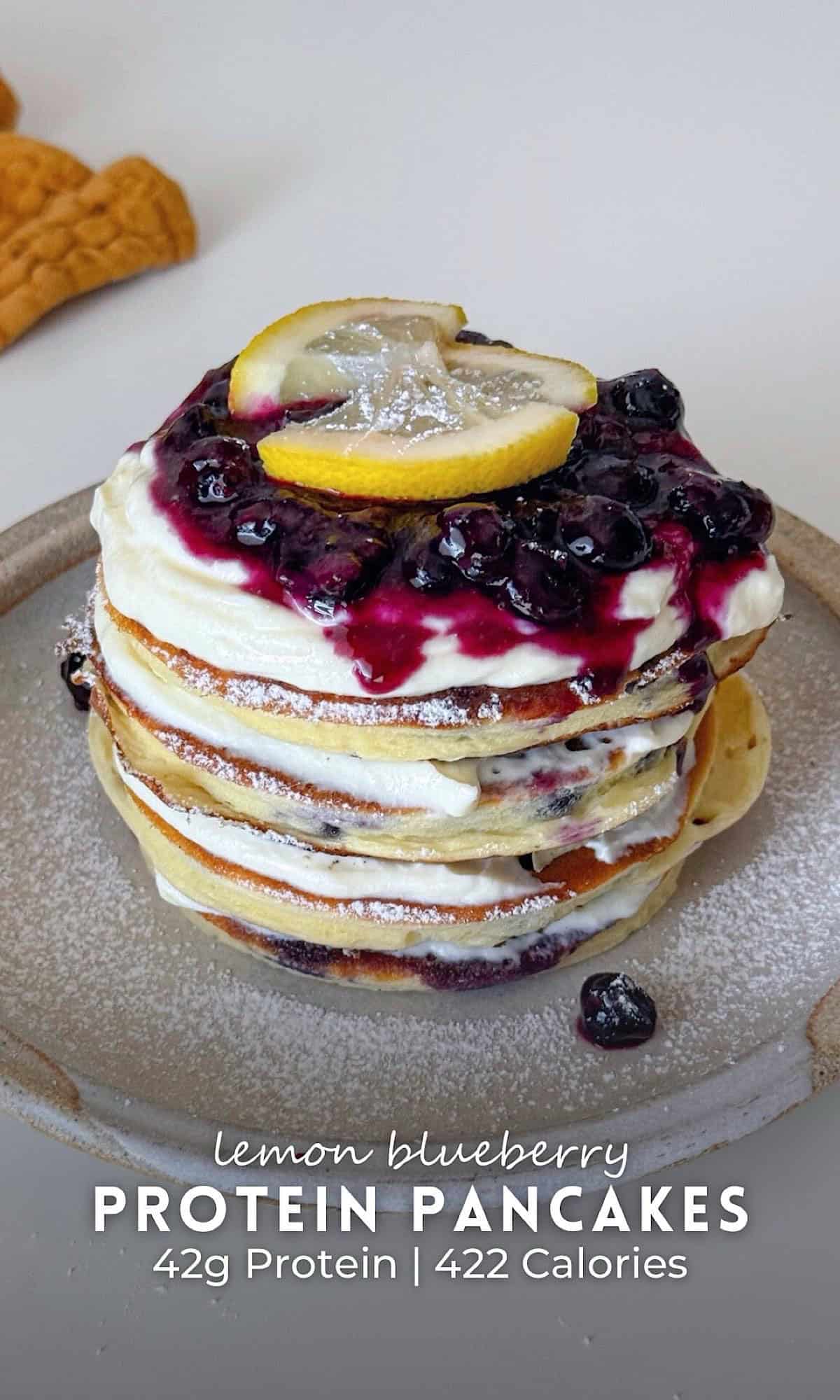 Lemon Blueberry Protein Pancakes 11 Stack of lemon blueberry protein pancakes layered with creamy lemon filling and topped with blueberry compote and lemon slices, served on a ceramic plate dusted with powdered sugar.