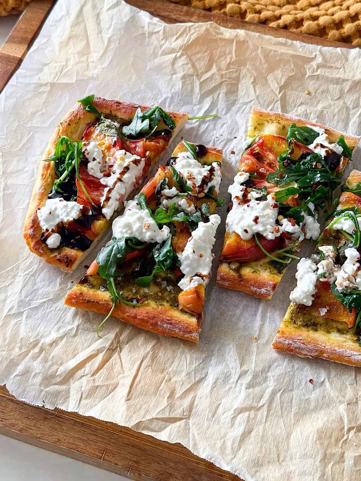 High Protein Grilled Peach Flatbread 1 Grilled peach flatbread topped with cottage cheese, arugula, heirloom tomatoes, pesto, balsamic glaze, and crushed red pepper, sliced into rectangular pieces on parchment paper.