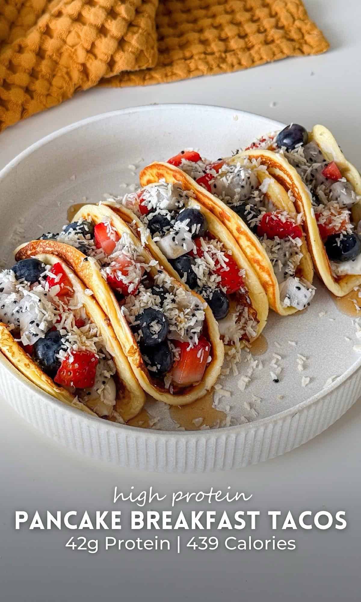 Fruit filled pancake breakfast tacos lined up in a white bowl, stuffed with fresh strawberries, blueberries, dragon fruit, and shredded coconut, drizzled with syrup for a colorful and refreshing summer brunch.