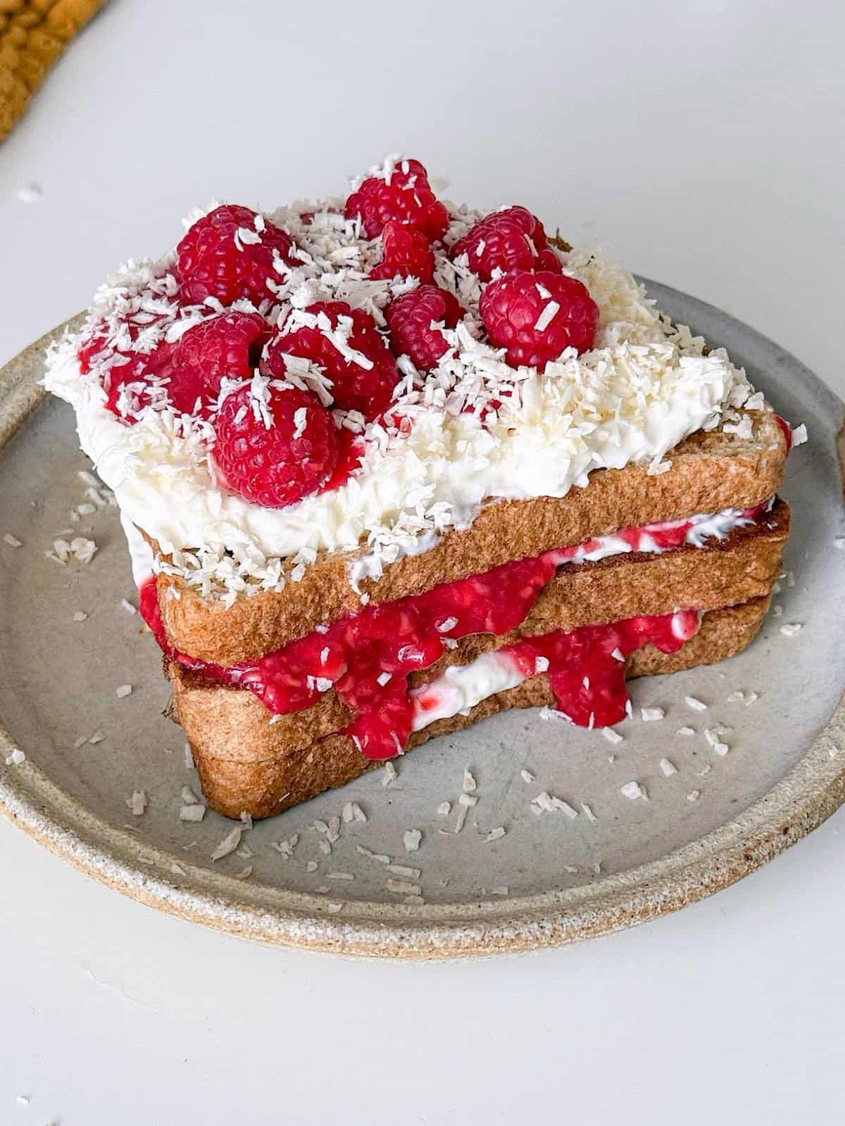 Raspberry Coconut Protein French Toast 1 Raspberry coconut protein French toast layered with creamy yogurt and raspberry compote, topped with fresh raspberries and shredded coconut on a neutral ceramic plate.