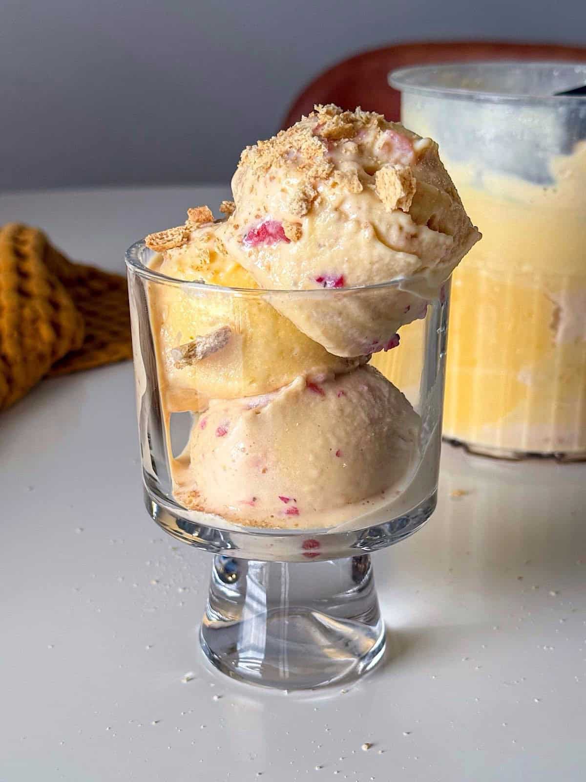 Strawberry Shortcake Custard Ice Cream 1 Strawberry shortcake custard ice cream scoops stacked in a clear glass, showing a creamy texture with visible strawberry pieces and crushed graham cracker topping. A Ninja Creami pint container is visible in the background.