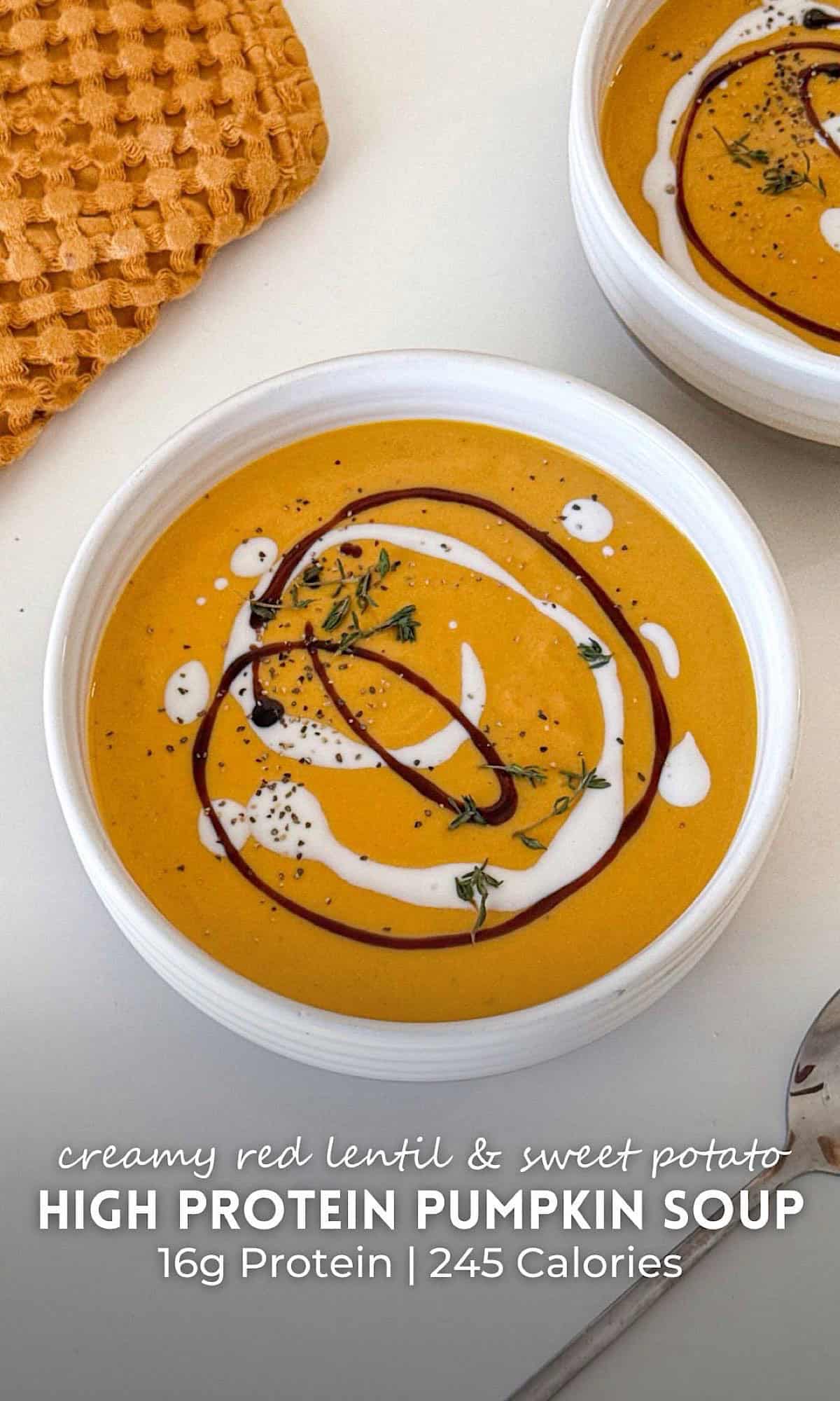 Creamy High Protein Pumpkin Soup 11 Two bowls of creamy high protein pumpkin soup drizzled with yogurt and balsamic glaze, topped with fresh thyme and black pepper, served on a white table beside a silver spoon and a textured orange cloth.
