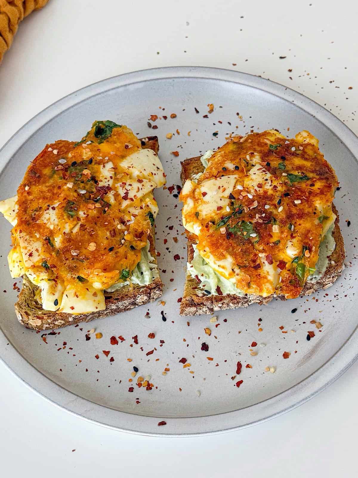 Crispy Egg Salad Avocado Toast 1 Two slices of crispy egg salad avocado toast topped with golden brown eggs, mashed avocado, and red pepper flakes on a light gray plate.