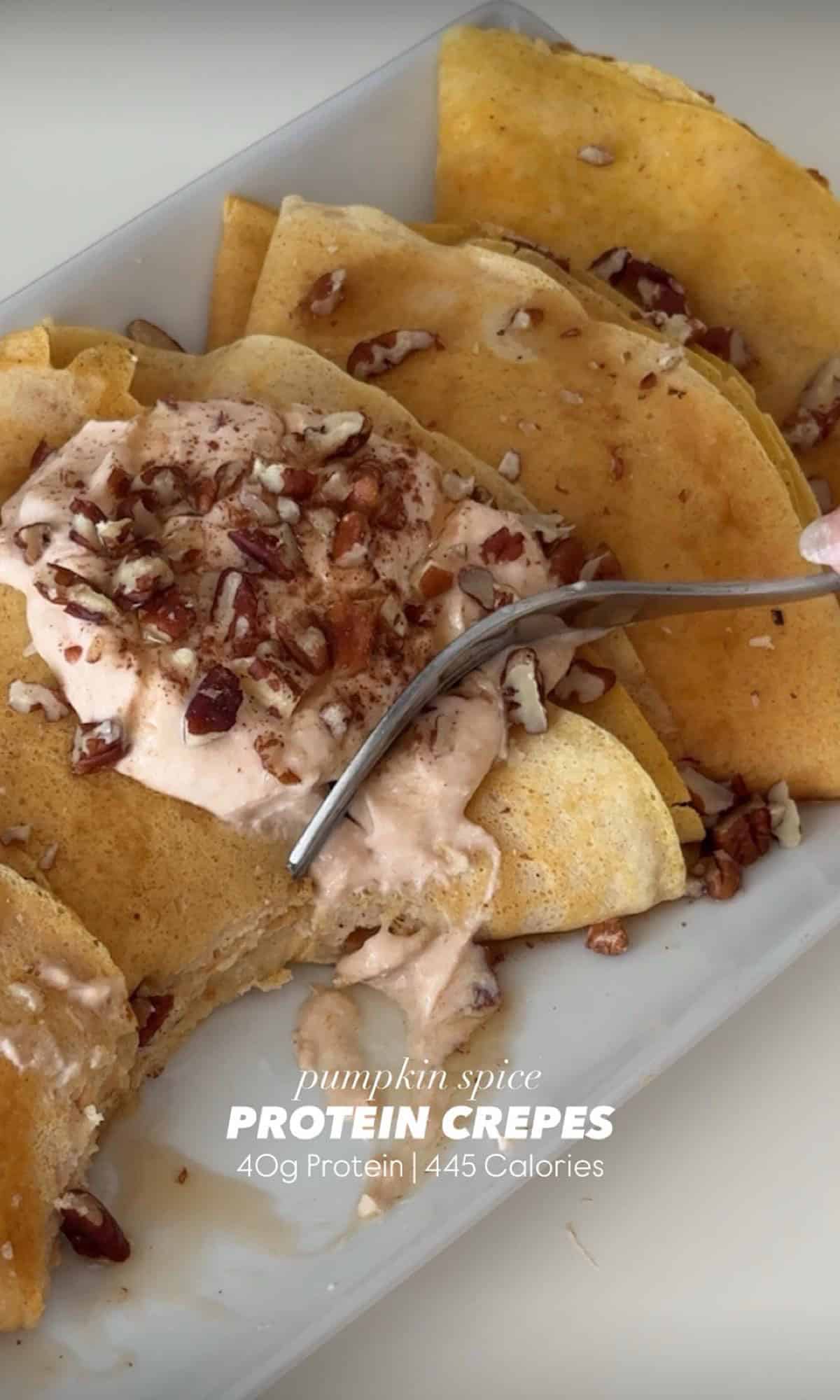 A rectangular plate with folded pumpkin pie protein crepes topped with a dollop of spiced yogurt, chopped pecans, and a drizzle of syrup. A warm-toned cloth napkin is placed in the background.