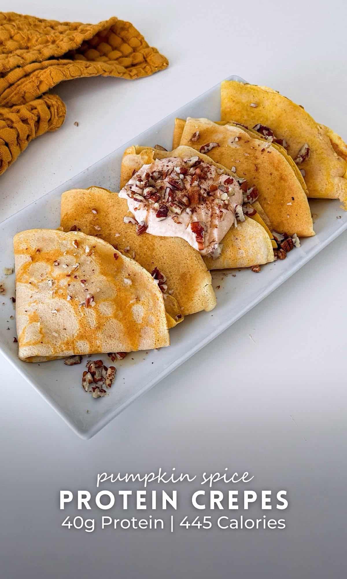 A rectangular plate with folded pumpkin pie protein crepes topped with a dollop of spiced yogurt, chopped pecans, and a drizzle of syrup. A warm-toned cloth napkin is placed in the background.