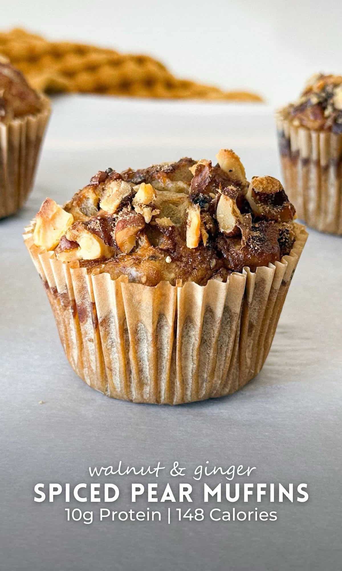 Freshly baked walnut & ginger spiced pear muffins with golden tops and crunchy walnut crumble, displayed on parchment paper with a cozy orange kitchen towel in the background.