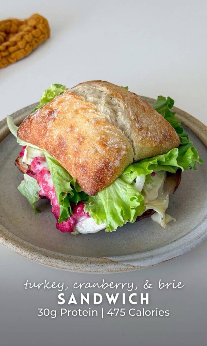 A hearty veggie turkey, cranberry, & brie sandwich stacked on a rustic ciabatta roll, filled with leafy greens, creamy melted brie, and a vibrant cranberry spread. The sandwich sits on a speckled ceramic plate over a clean white surface, with a textured mustard cloth in the background adding warmth to the scene.
