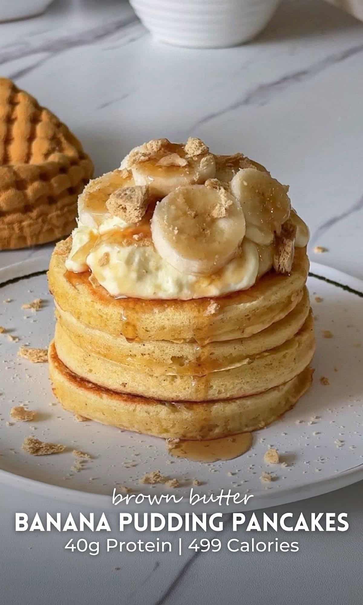 Brown Butter Banana Pudding Pancakes 10 Brown butter banana pudding pancakes stacked on a white plate, featuring fluffy golden pancakes topped with creamy banana pudding, fresh banana slices, crushed vanilla wafers, and a drizzle of syrup. The stack sits on a marble surface with a textured mustard cloth and kitchen decor softly blurred in the background.