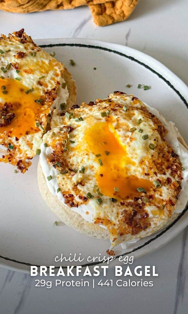 Chili crisp egg breakfast bagel served open-faced on a plate, featuring toasted bagel halves topped with a creamy spread and crispy-edged fried eggs with jammy yolks. The eggs are seasoned with chili crisp and garnished with chopped chives, with the rich yolk spilling over the edges onto the bagel.