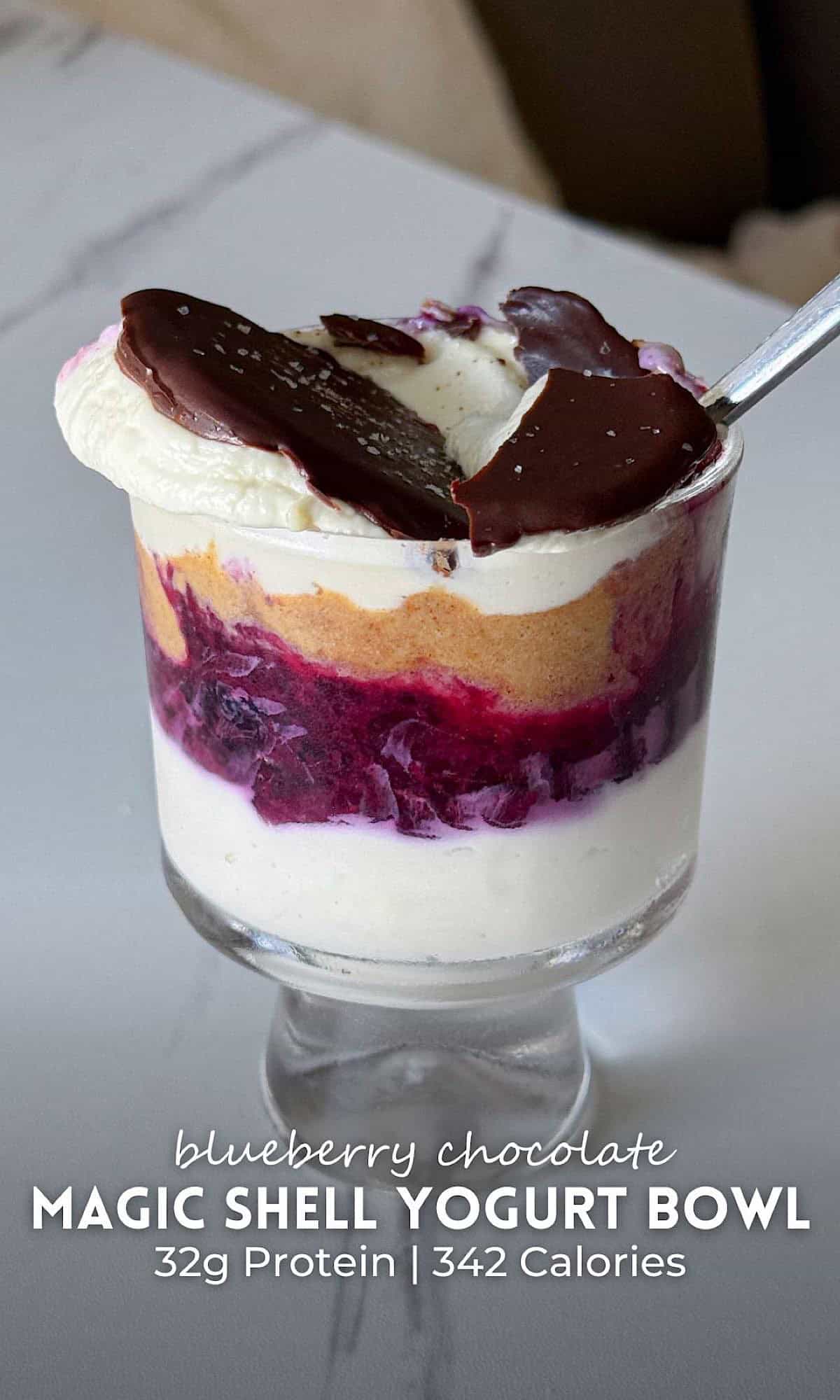 Alt text:
Chocolate magic shell yogurt bowl layered in a clear glass, featuring creamy yogurt, a vibrant berry compote layer, and a smooth nutty spread, topped with a hardened chocolate shell cracked open by a spoon. The layered dessert sits on a marble surface, highlighting the contrast between the creamy yogurt and glossy chocolate topping.