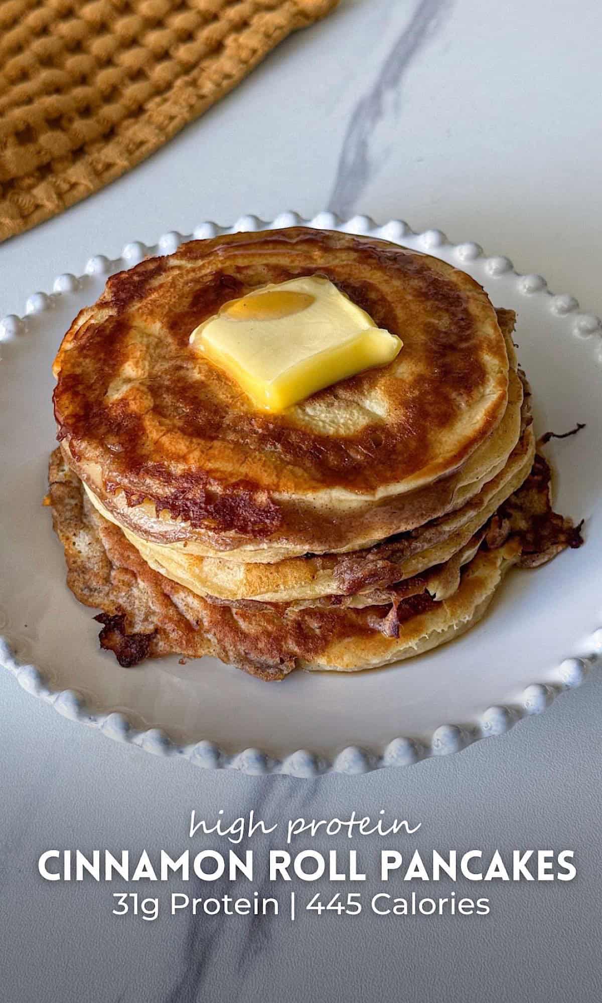 Stack of cinnamon roll pancakes on a white plate, golden and fluffy with swirled cinnamon layers, topped with a melting pat of butter.
