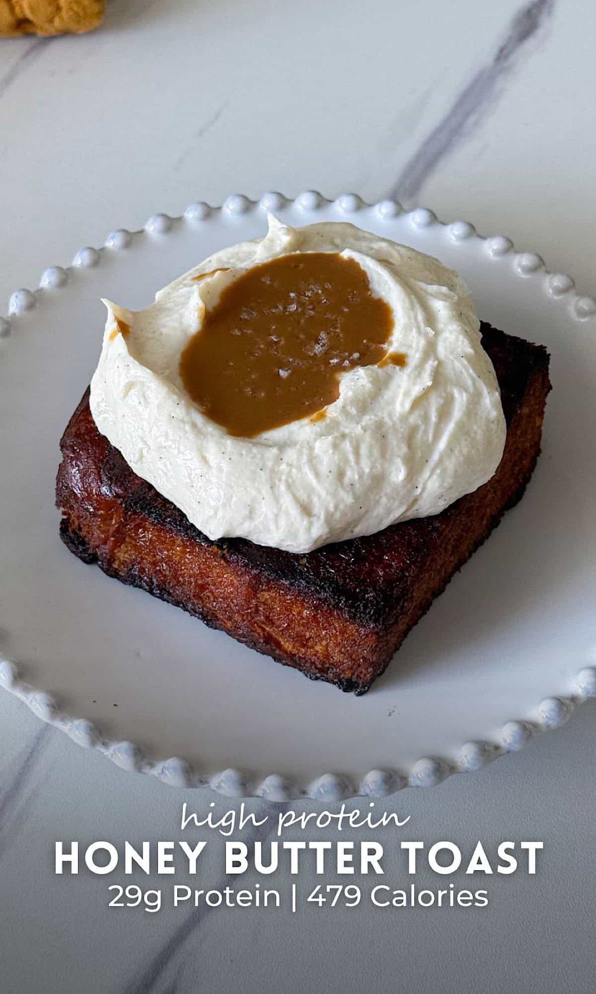 High protein honey butter toast served on a white beaded plate, featuring a thick golden slice of toast topped with a fluffy whipped cream mixture and a drizzle of honey butter in the center. The toast has crisp caramelized edges and sits on a marble surface with a textured mustard cloth in the background.