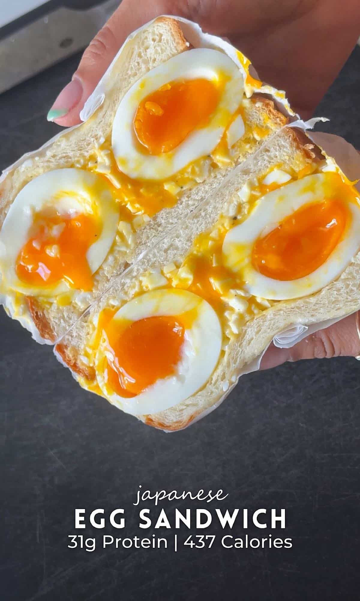 Japanese egg sandwich held in hand, made with soft white bread filled with creamy egg salad and halved soft-boiled eggs with jammy yolks oozing out.