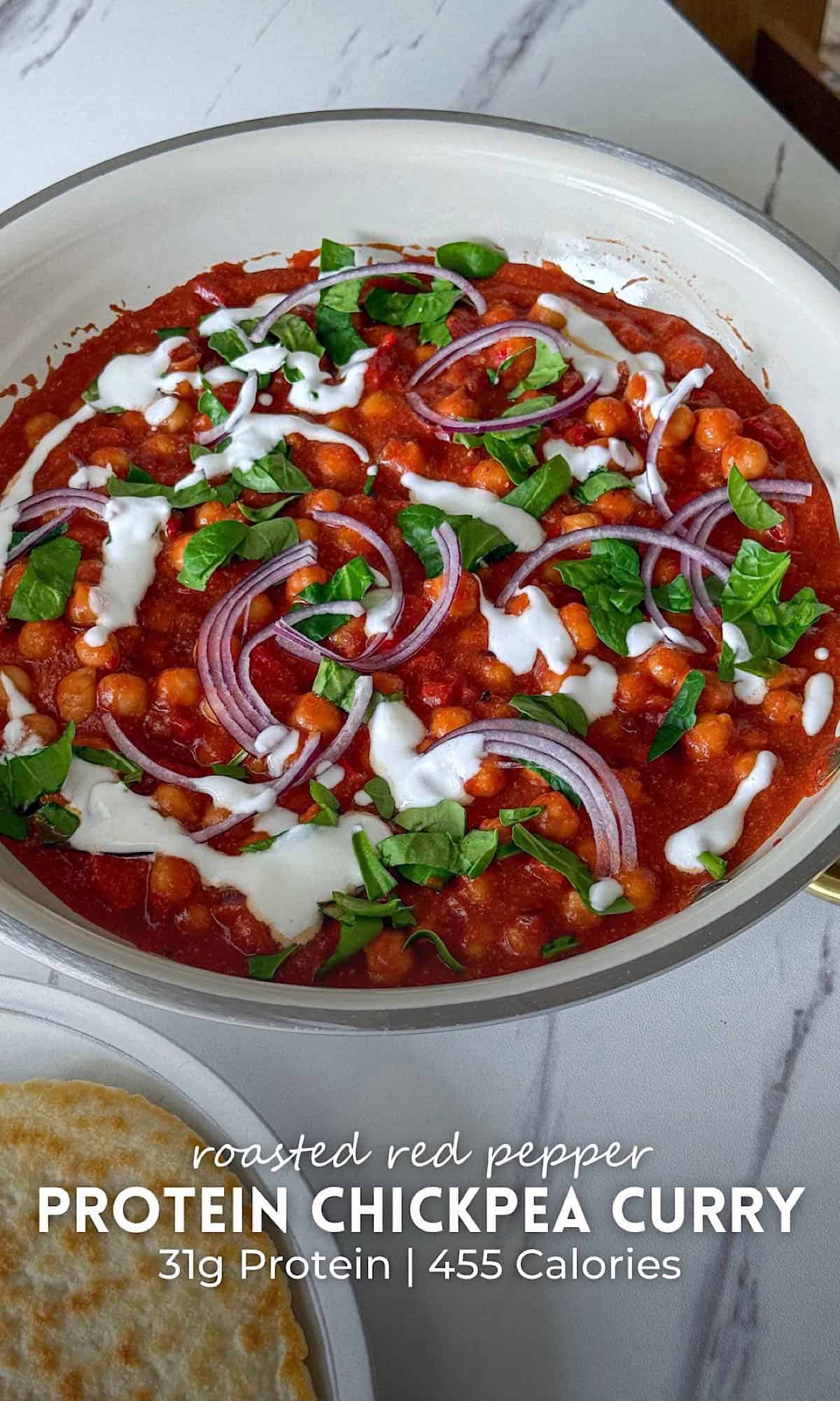 Roasted red pepper chickpea curry served in a white skillet, featuring chickpeas simmered in a rich roasted red pepper tomato sauce and topped with fresh greens, thinly sliced red onion, and a drizzle of creamy sauce. The curry sits on a marble surface with toasted flatbread on a plate beside it.