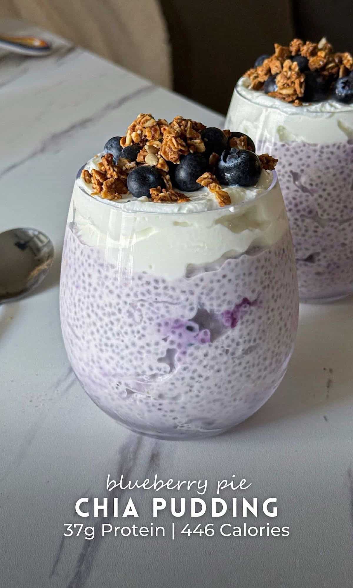 Two glasses of blueberry pie chia pudding layered with creamy yogurt, topped with fresh blueberries and crunchy granola, served on a marble countertop.