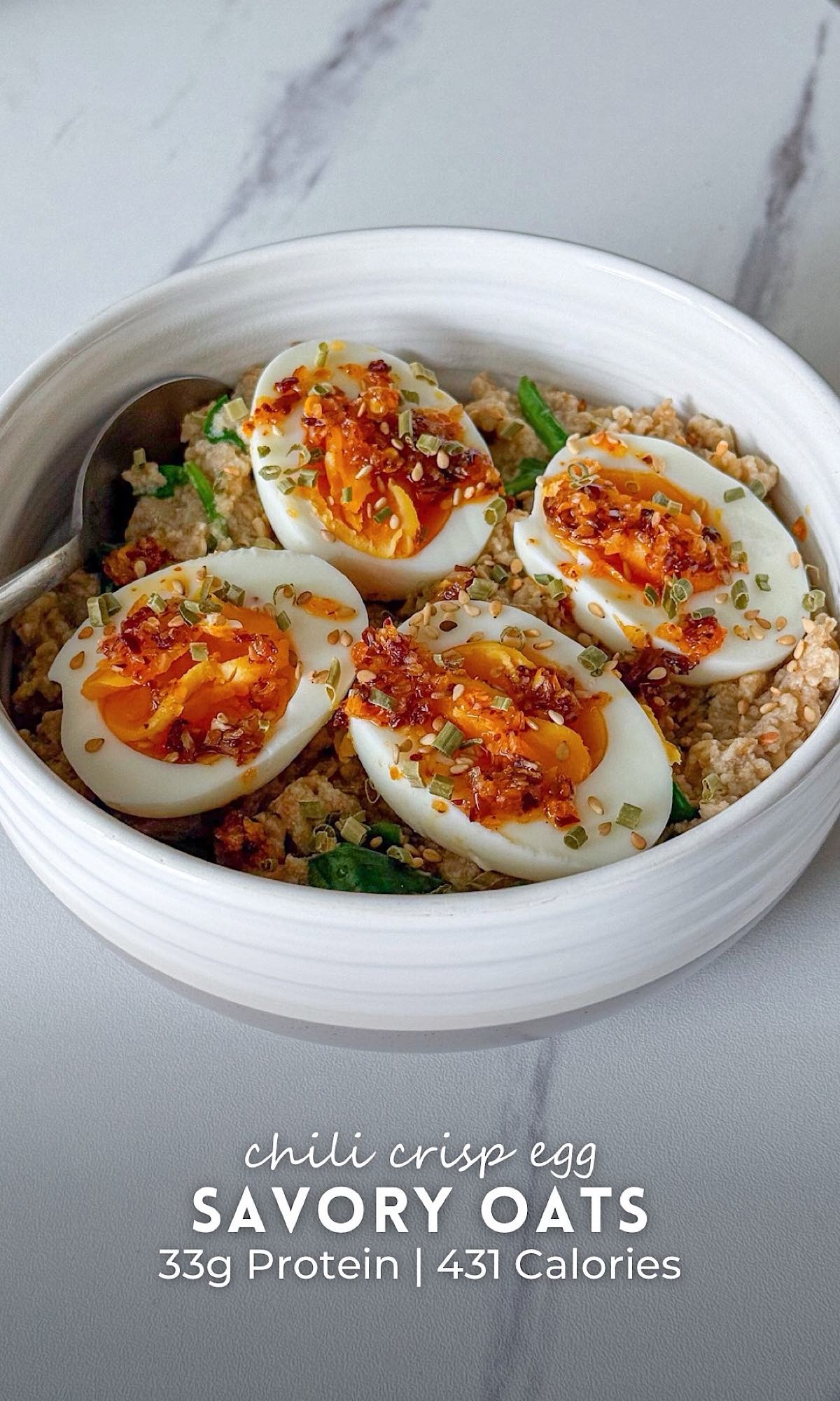 Chili crisp egg savory oats in a white bowl, topped with soft boiled eggs, chili crisp, sesame seeds, and chopped herbs, served with a spoon on a marble countertop.