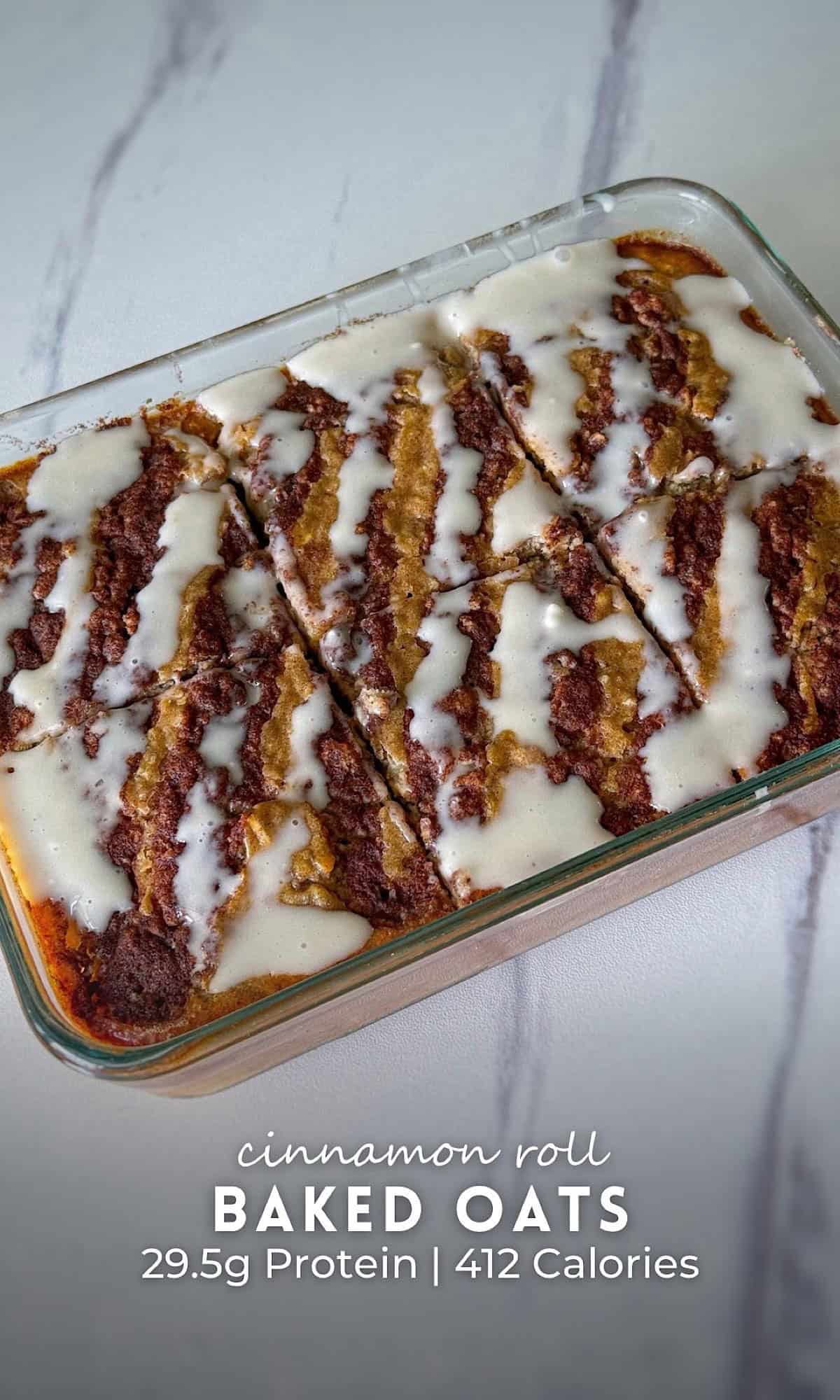 Cinnamon roll baked oats sliced in a glass baking dish, swirled with cinnamon filling and topped with a creamy vanilla glaze, served on a marble surface.