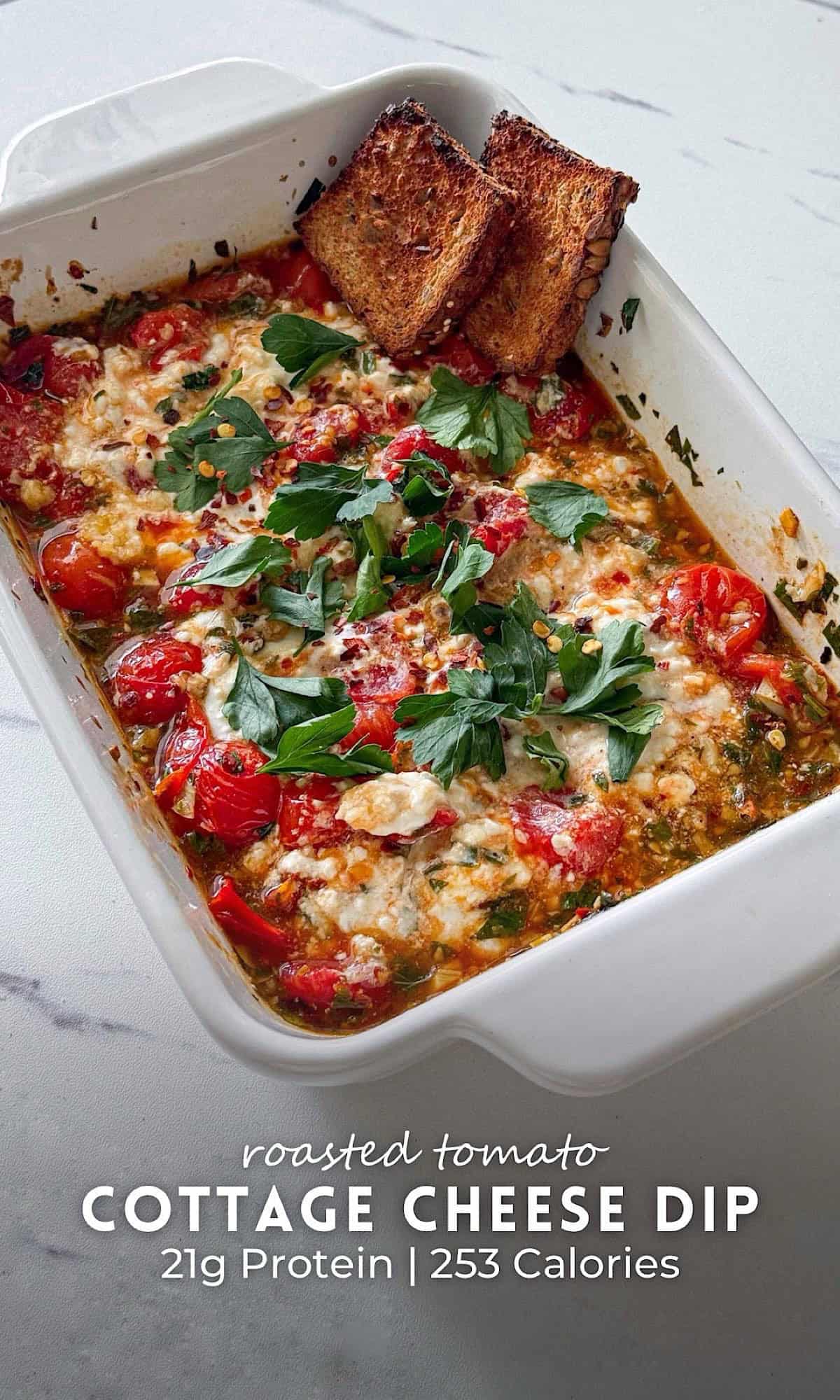 Roasted tomato cottage cheese dip baked in a white dish, topped with fresh cilantro, red pepper flakes, and melted cheese, served with toasted bread slices on the side.