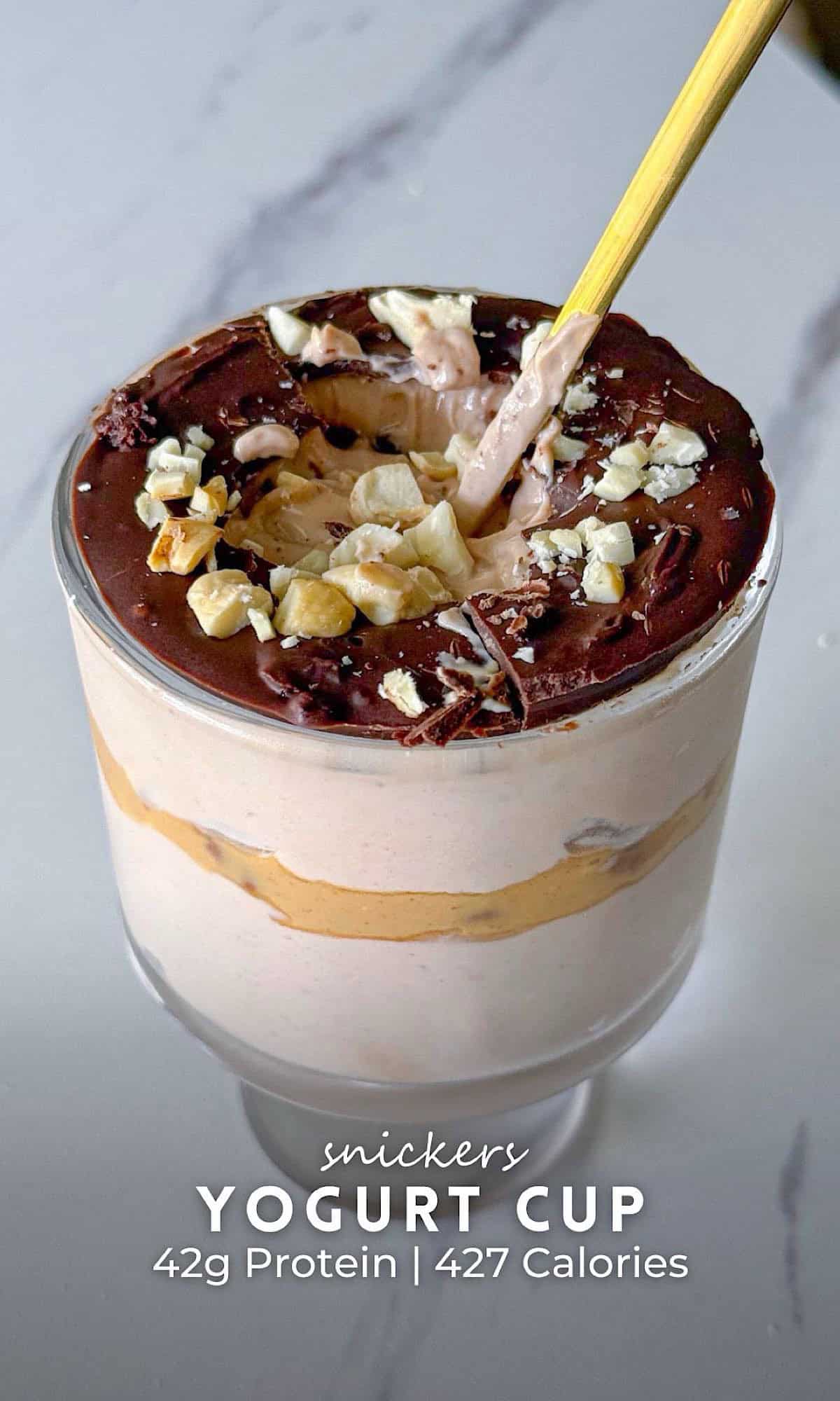 Snickers yogurt cup layered with creamy yogurt and a peanut butter swirl, topped with a chocolate magic shell, chopped nuts, and chocolate shavings, with a spoon cracking through the shell.
