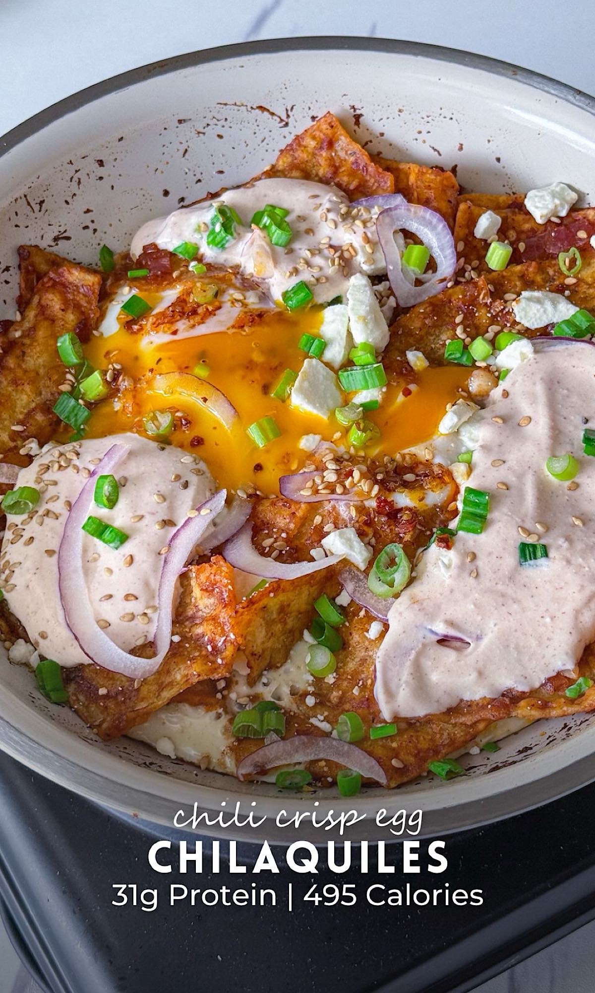 Chili Crisp Egg Chilaquiles 9 Chili crisp egg chilaquiles made with crispy tortilla chips in a creamy red sauce, topped with jammy eggs, crumbled cheese, green onions, red onion, sesame seeds, and a spicy creamy drizzle in a white nonstick pan.