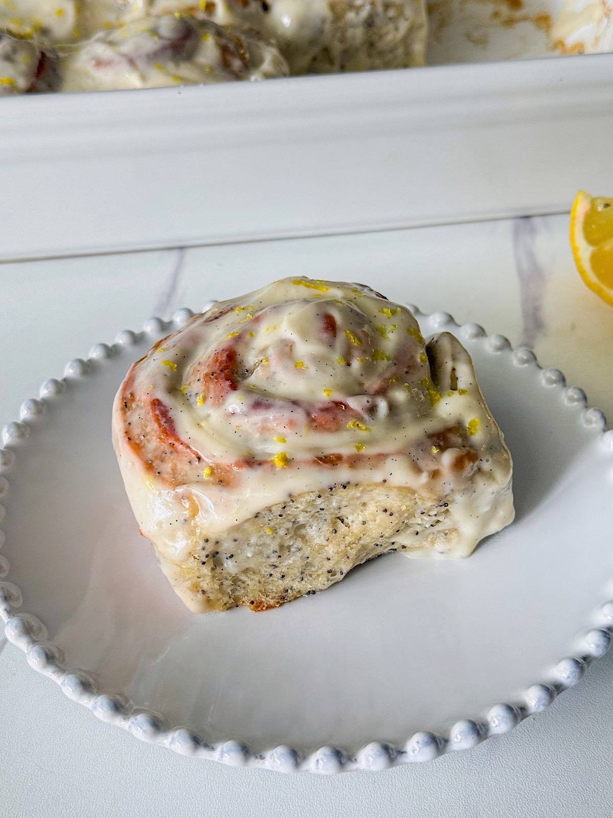 Lemon Poppy Seed Rolls 1 Soft and fluffy lemon poppy seed rolls covered in a thick lemon cream cheese glaze, with visible poppy seeds and fresh lemon zest on top, served on a white beaded plate.