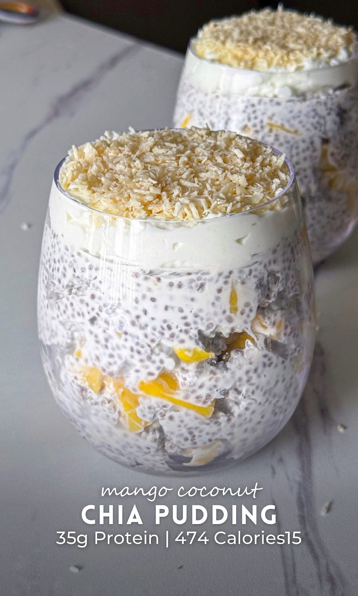 Two glasses of mango coconut chia pudding layered with fresh mango chunks and creamy yogurt, topped with shredded coconut on a marble countertop.