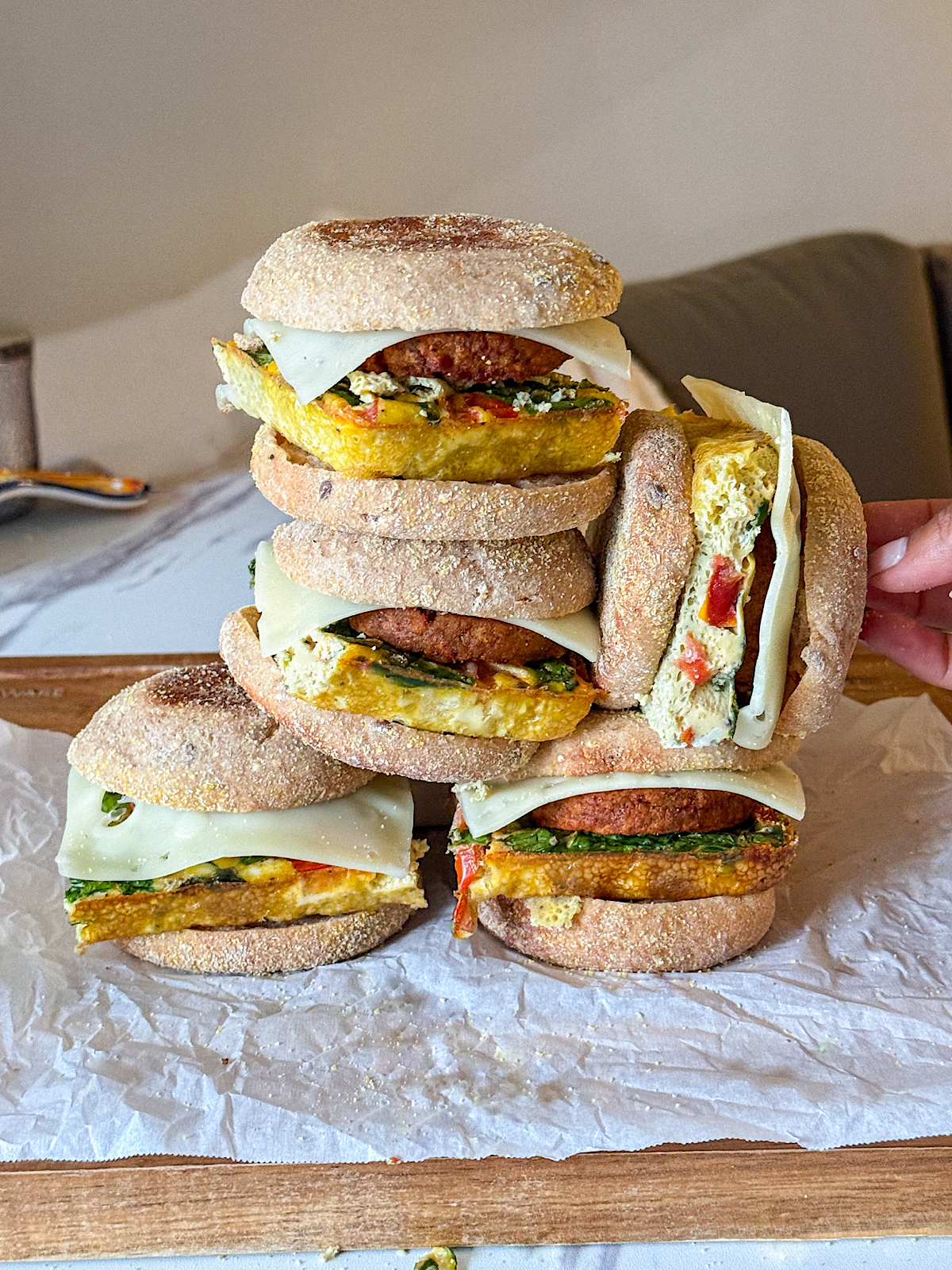 Meal Prep Breakfast Sandwiches 1 Stack of meal prep breakfast sandwiches made with English muffins, fluffy veggie egg patties, vegetarian sausage, spinach, and melted cheese on a wooden board with one sandwich cut open to show the filling.