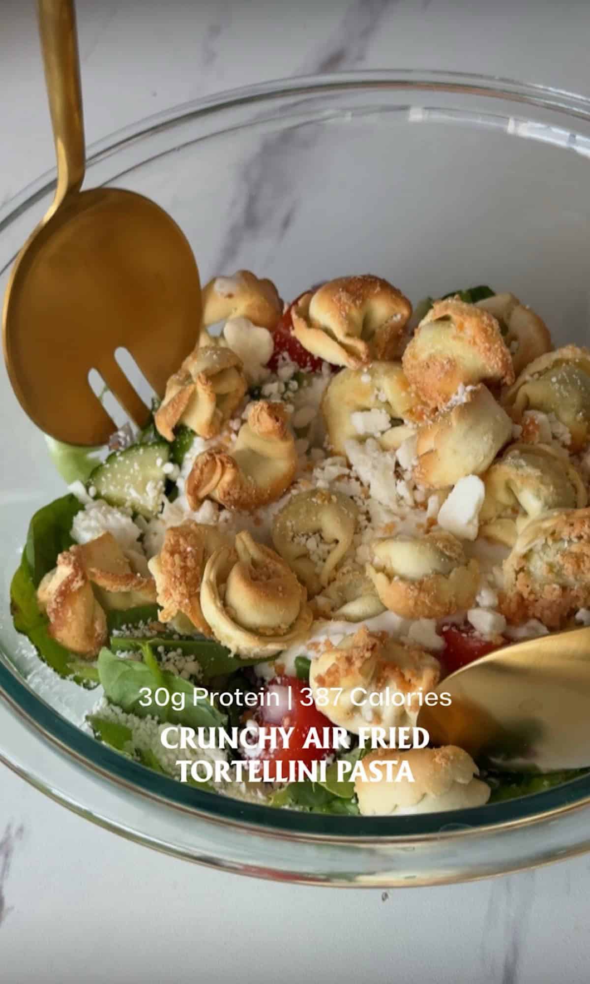Crunchy air fried tortellini salad made with crispy golden cheese tortellini served over fresh romaine lettuce, cucumbers, cherry tomatoes, and red onion in a clear glass bowl. The salad is topped with crumbled feta and grated parmesan, creating a colorful, protein-packed lunch with plenty of crispy texture.