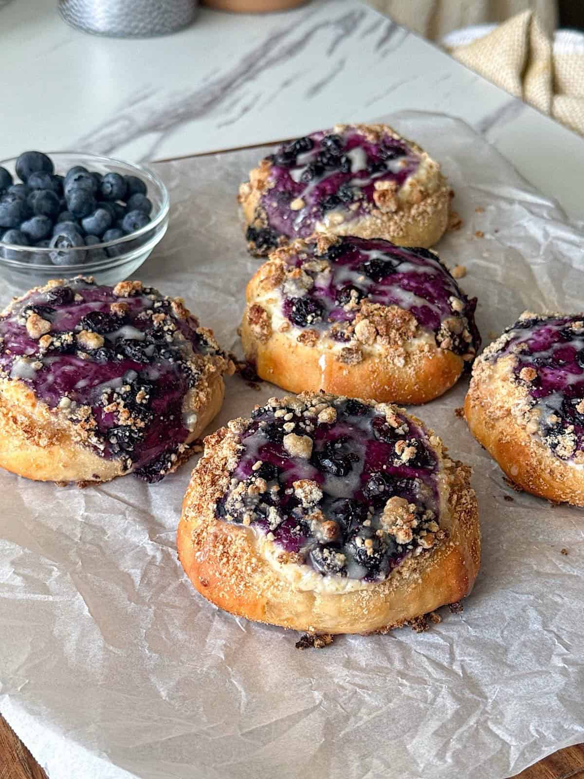Protein Blueberry Cheesecake Buns 1 Protein blueberry cheesecake buns baked until golden brown and topped with a creamy cheesecake filling, juicy blueberries, buttery crumble, and a light vanilla glaze. The buns are arranged on parchment paper with a bowl of fresh blueberries nearby, making a high protein breakfast or dessert with a soft, bakery-style texture.