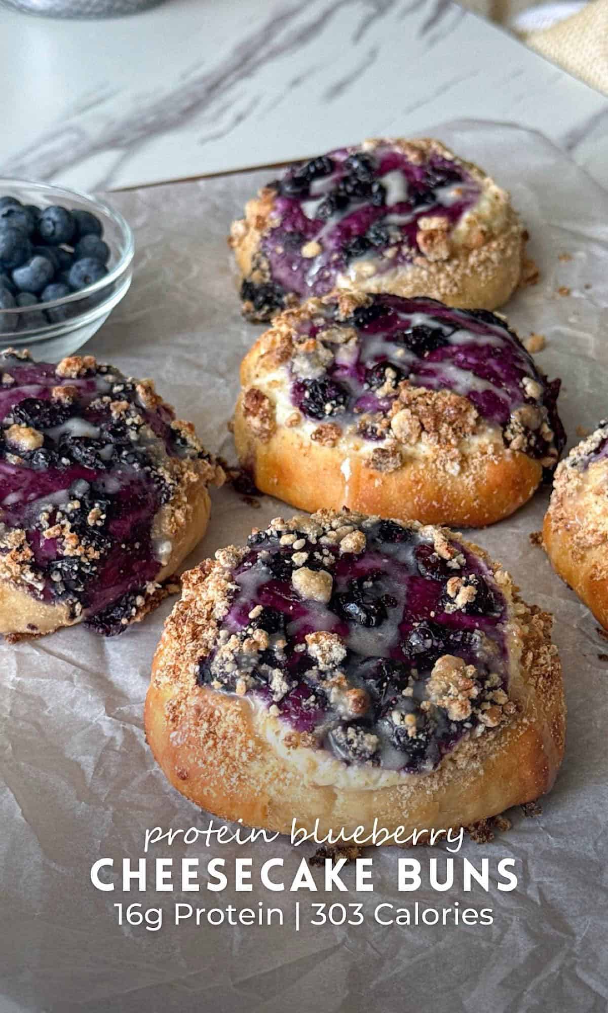 Protein Blueberry Cheesecake Buns 11 Protein blueberry cheesecake buns baked until golden brown and topped with a creamy cheesecake filling, juicy blueberries, buttery crumble, and a light vanilla glaze. The buns are arranged on parchment paper with a bowl of fresh blueberries nearby, making a high protein breakfast or dessert with a soft, bakery-style texture.