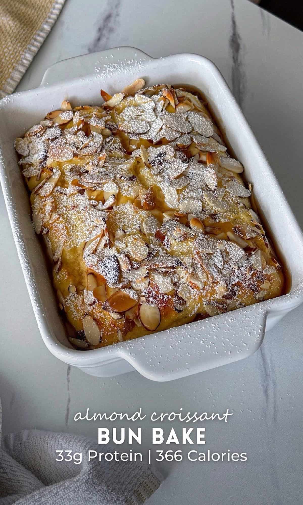 Almond croissant bun bake baked until golden brown in a white ceramic baking dish, topped with toasted sliced almonds and a generous dusting of powdered sugar. This high protein almond croissant bun bake has a soft, fluffy interior with crisp toasted almonds and a buttery bakery-inspired finish.