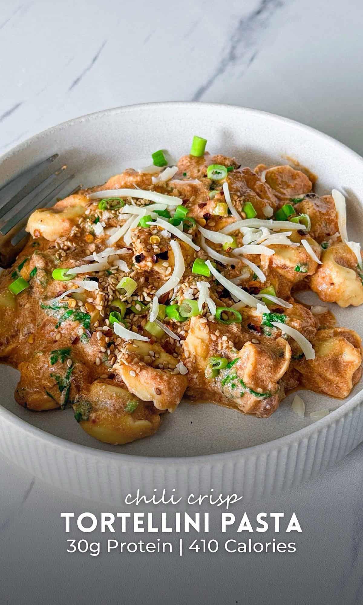 Chili Crisp Tortellini Pasta 10 Chili crisp tortellini pasta tossed in a creamy, spicy chili crisp sauce and served in a shallow white bowl. The cheese tortellini is finished with shredded parmesan, sliced green onions, sesame seeds, and red pepper flakes, creating a rich, high protein vegetarian pasta with bold flavor and a creamy texture.