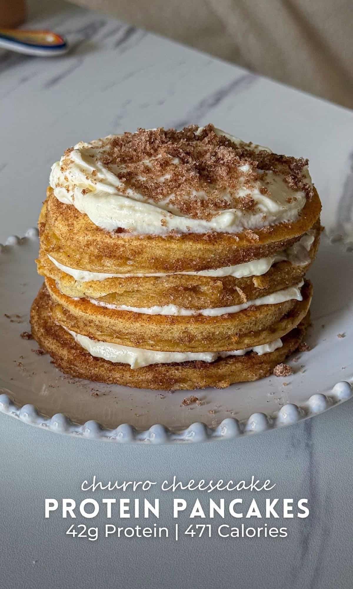 Churro Cheesecake Protein Pancakes 11 Stack of churro cheesecake protein pancakes layered with creamy cheesecake filling, topped with more filling and a dusting of cinnamon sugar, served on a white beaded plate.