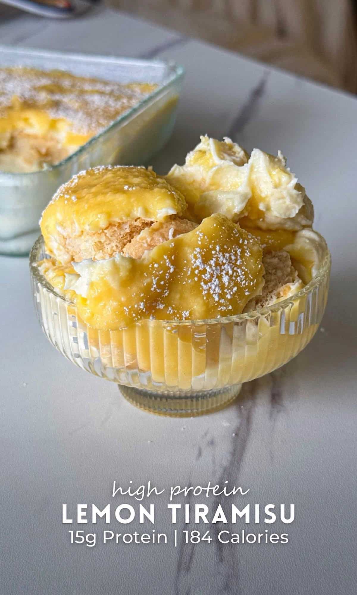 High Protein Lemon Tiramisu with Greek Yogurt 10 High protein lemon tiramisu layered with lemon curd, creamy Greek yogurt filling, and ladyfingers, served in a glass dessert bowl with whipped topping and powdered sugar, with the baking dish in the background.