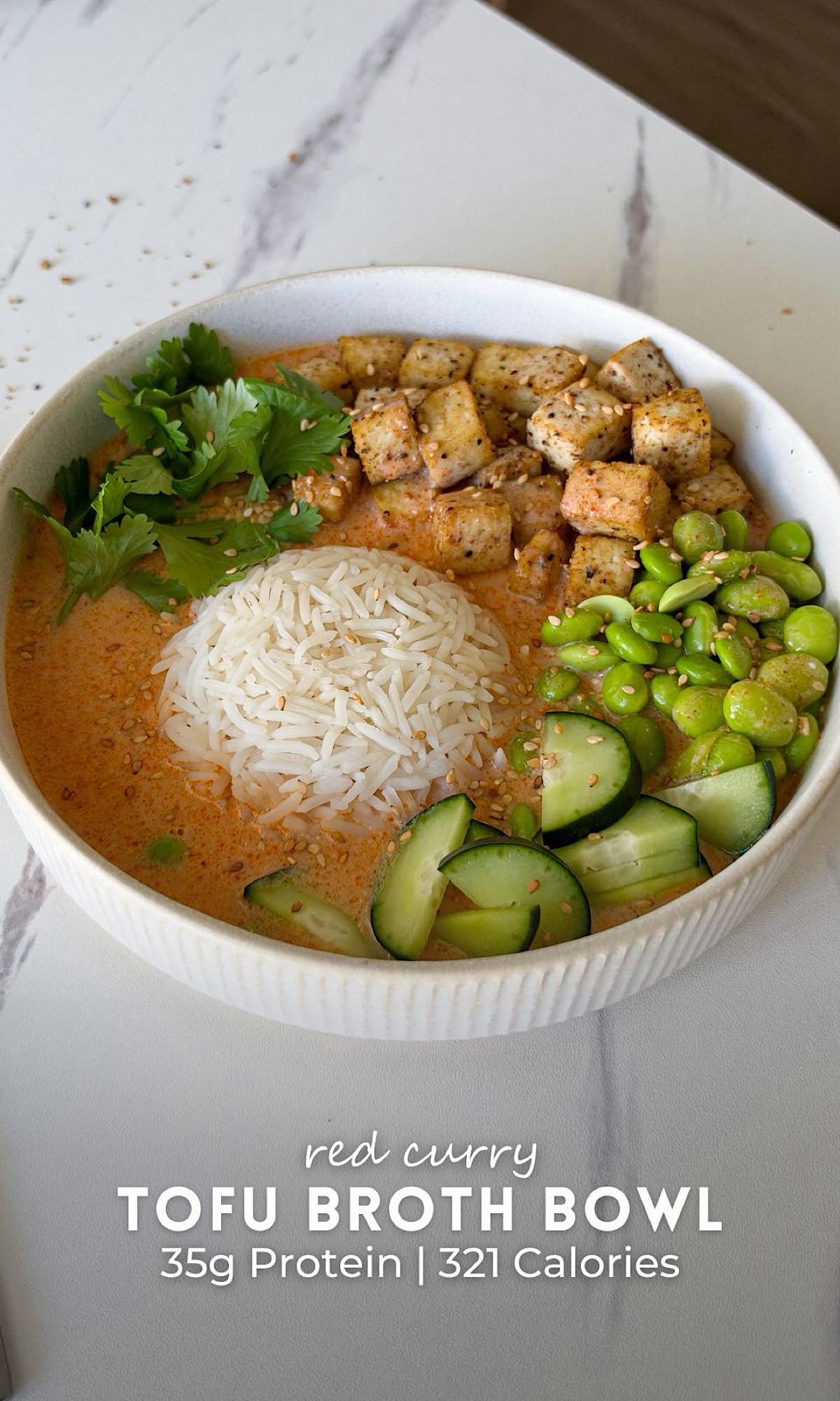 Red curry tofu broth bowl served in a white ceramic bowl with creamy orange broth, white rice, seasoned tofu cubes, edamame, sliced cucumbers, fresh cilantro, and sesame seeds.